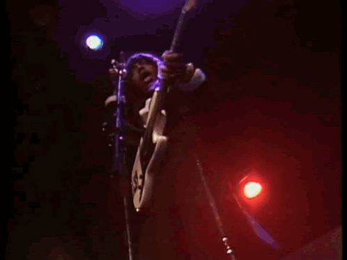 A Man Playing A Guitar And Singing Into A Microphone On A Stage GIF