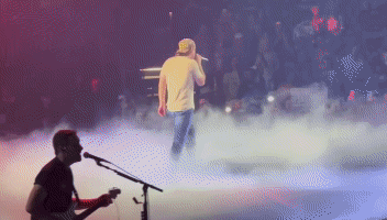 A Man Playing A Guitar And Singing Into A Microphone On A Stage GIF