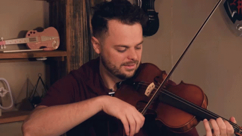 A Man Playing A Violin In Front Of A Shelf With A Pink Guitar That Says ' Elvis ' On It GIF
