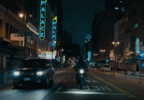 A Man Riding A Motorcycle Down A City Street With A Neon Sign That Says Palace GIF