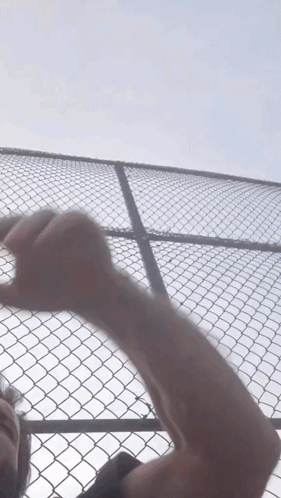 A Man 'S Arm Is Visible Behind A Chain Link Fence Against A Blue Sky GIF