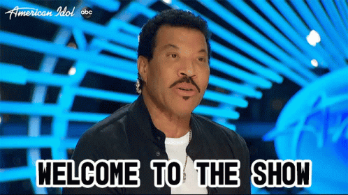 A Man Says Welcome To The Show While Sitting In Front Of A Blue Background GIF