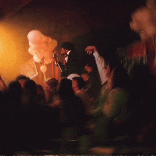 A Man Singing Into A Microphone In Front Of A Crowd With Balloons In The Background GIF