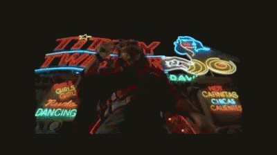 A Man Singing Into A Microphone In Front Of A Lit Up Sign That Says Titty Twist GIF