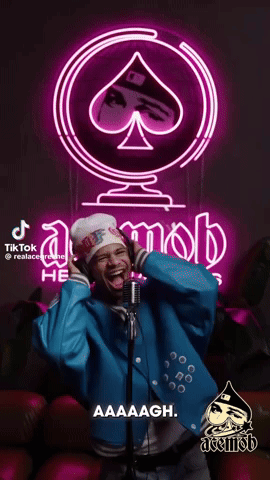A Man Singing Into A Microphone In Front Of A Neon Sign That Says Acemob GIF