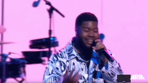 A Man Singing Into A Microphone In Front Of A Pink Background With The Word Aria On It GIF