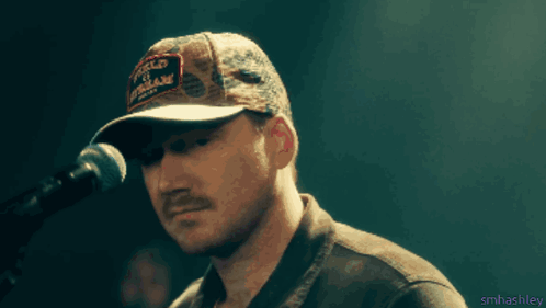 A Man Singing Into A Microphone Wearing A Hat That Says " World Of Motorcycles " GIF
