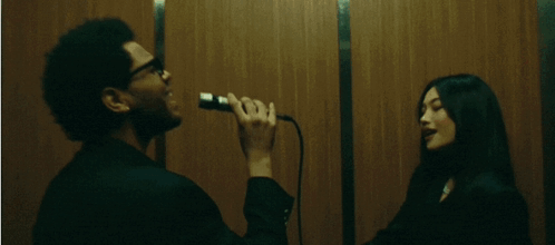 A Man Singing Into A Microphone While A Woman Looks On GIF