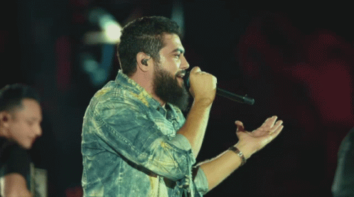A Man Singing Into A Microphone While Another Man Plays A Guitar In Front Of A Crowd GIF