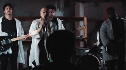 A Man Singing Into A Microphone While Playing A Guitar And A Drum Set GIF