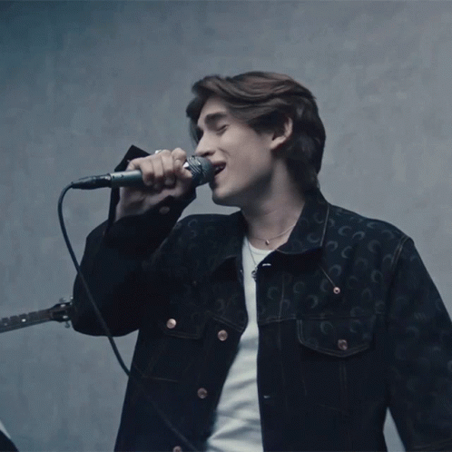 A Man Singing Into A Microphone While Wearing A Black Jacket With The Letter C On It GIF