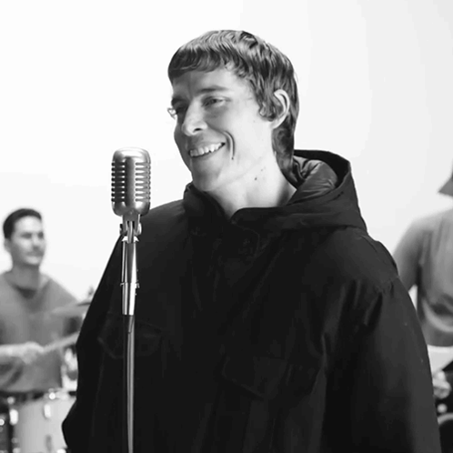 A Man Singing Into A Microphone With A Band Behind Him GIF