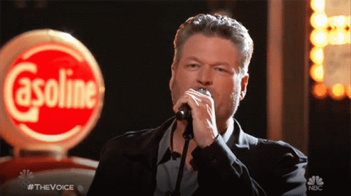 A Man Singing Into A Microphone With A Gasoline Sign In The Background GIF