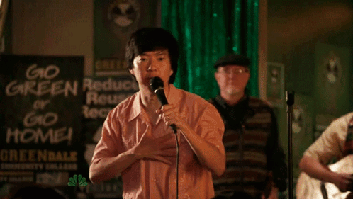 A Man Singing Into A Microphone With A Sign Behind Him That Says Go Green Or Go Home GIF