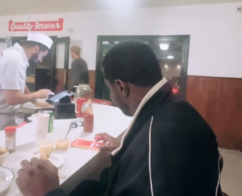 A Man Sits At A Counter In Front Of A Sign That Says " Quality Forever " GIF
