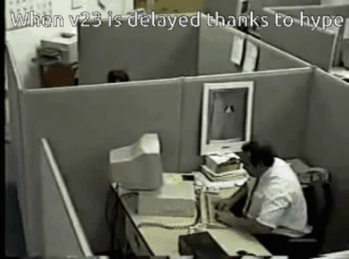 A Man Sits At A Desk In A Cubicle With The Words When V23 Is Delayed Thanks To Hype GIF
