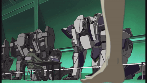 A Man Sits At A Desk In Front Of A Row Of Robots And A Person Standing Next To Them GIF