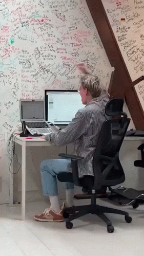 A Man Sits At A Desk With A Laptop In Front Of A Wall That Has Graffiti On It Including The Name Camelle GIF