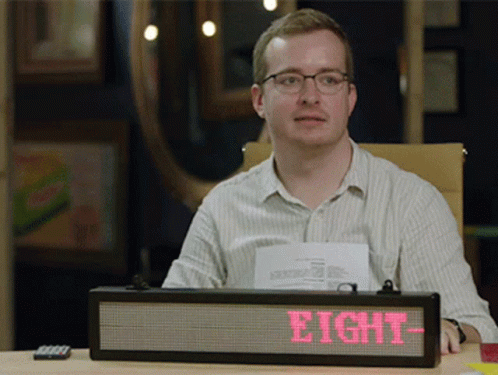 A Man Sits At A Desk With A Sign That Says Eight On It GIF