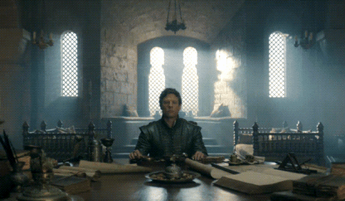 A Man Sits At A Table In A Dark Room With A Sword In His Hand GIF