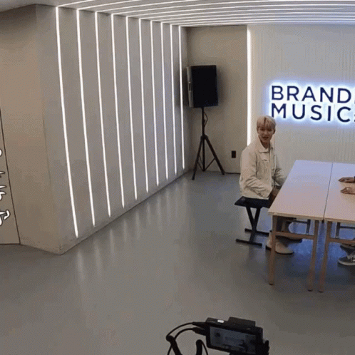 A Man Sits At A Table In A Room With A Sign That Says Brand Music GIF