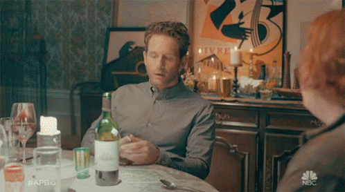 A Man Sits At A Table With A Bottle Of Wine In Front Of A Poster That Says Livre GIF