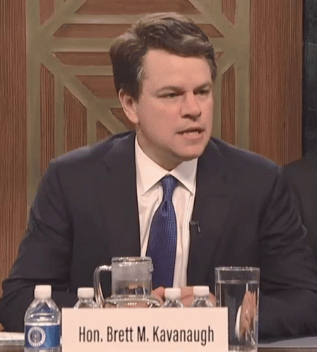 A Man Sits At A Table With A Sign That Says Hon. Brett M. Kavanugh GIF