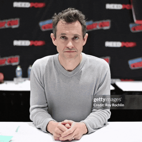 A Man Sits At A Table With His Hands Folded In Front Of A Wall That Says Reedpop GIF