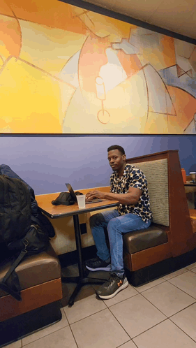 A Man Sits In A Booth At A Table With A Laptop On It GIF