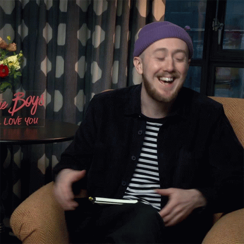 A Man Sits In A Chair In Front Of A Sign That Says Boys Love You GIF
