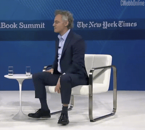 A Man Sits In A Chair In Front Of A Sign That Says The New York Times Book Summit GIF