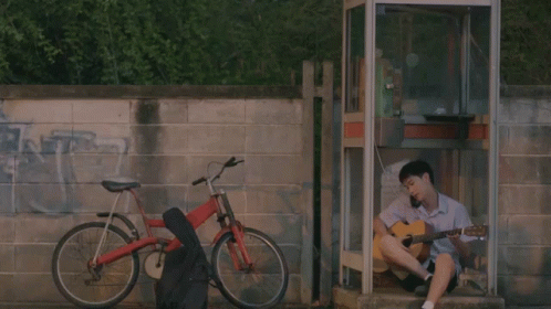 A Man Sits In A Phone Booth Playing A Guitar Next To A Bike GIF