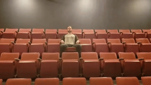 A Man Sits In A Row Of Red Seats In A Theater GIF