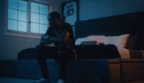 A Man Sits On A Bed In Front Of A Window With A Jersey On The Wall That Says 83 GIF