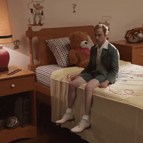 A Man Sits On A Bed With A Teddy Bear On It GIF
