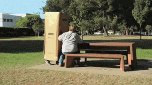 A Man Sits On A Bench In A Park While Carrying A Box GIF