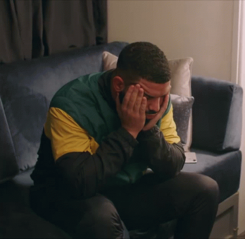 A Man Sits On A Blue Couch With His Hands On His Face GIF