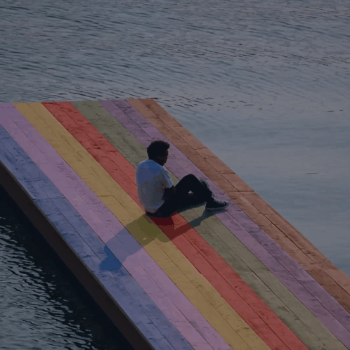 A Man Sits On A Colorful Wooden Dock Overlooking A Body Of Water GIF