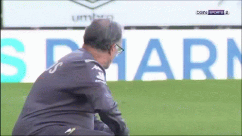 A Man Sits On A Soccer Field In Front Of An Ad For Bein Sports 2 GIF