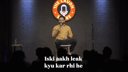 A Man Sits On A Stage In Front Of A Sign That Says " The Laugh Store " GIF