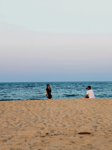 A Man Sits On The Beach While A Woman Stands On The Sand GIF
