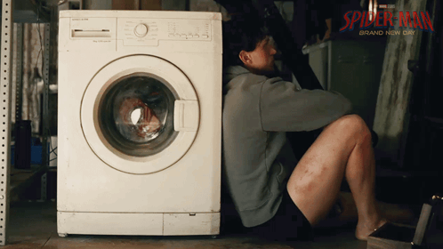 A Man Sits On The Floor Next To A Washing Machine That Says Spider-Man Brand Now On It GIF