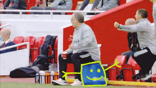 A Man Sits On The Sidelines Of A Soccer Game With A Smiley Face Drawn On A Blue Bag GIF