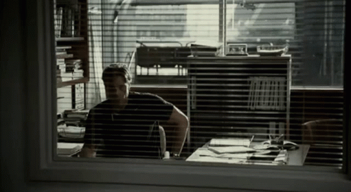 A Man Sitting At A Desk In Front Of A Window With Blinds GIF