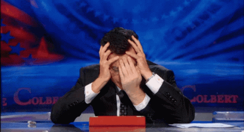 A Man Sitting At A Desk With His Hands On His Head In Front Of A Colbert Sign GIF