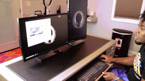 A Man Sitting At A Desk With Two Monitors And A Trash Can In The Background GIF