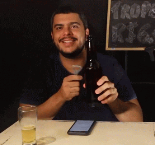 A Man Sitting At A Table Holding A Beer Bottle GIF