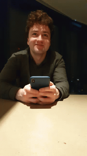 A Man Sitting At A Table Holding A Cell Phone In His Hands GIF