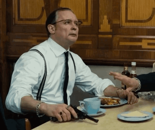 A Man Sitting At A Table Holding A Gun In His Hand GIF