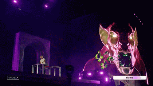 A Man Sitting At A Table In Front Of A Stage With A Sign That Says " Flume " On It GIF